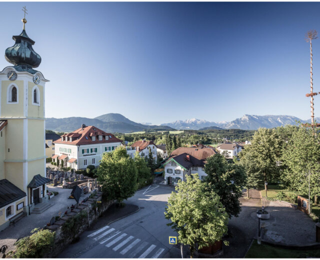 Ein malerisches österreichisches Dorf wie Elixhausen mit einer Kirche, Bäumen, Häusern und Bergen unter einem klaren Himmel.