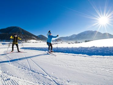 Zwei Personen genießen das Langlaufen auf einer verschneiten Strecke mit Bergen und einer strahlenden Sonne im Hintergrund.
