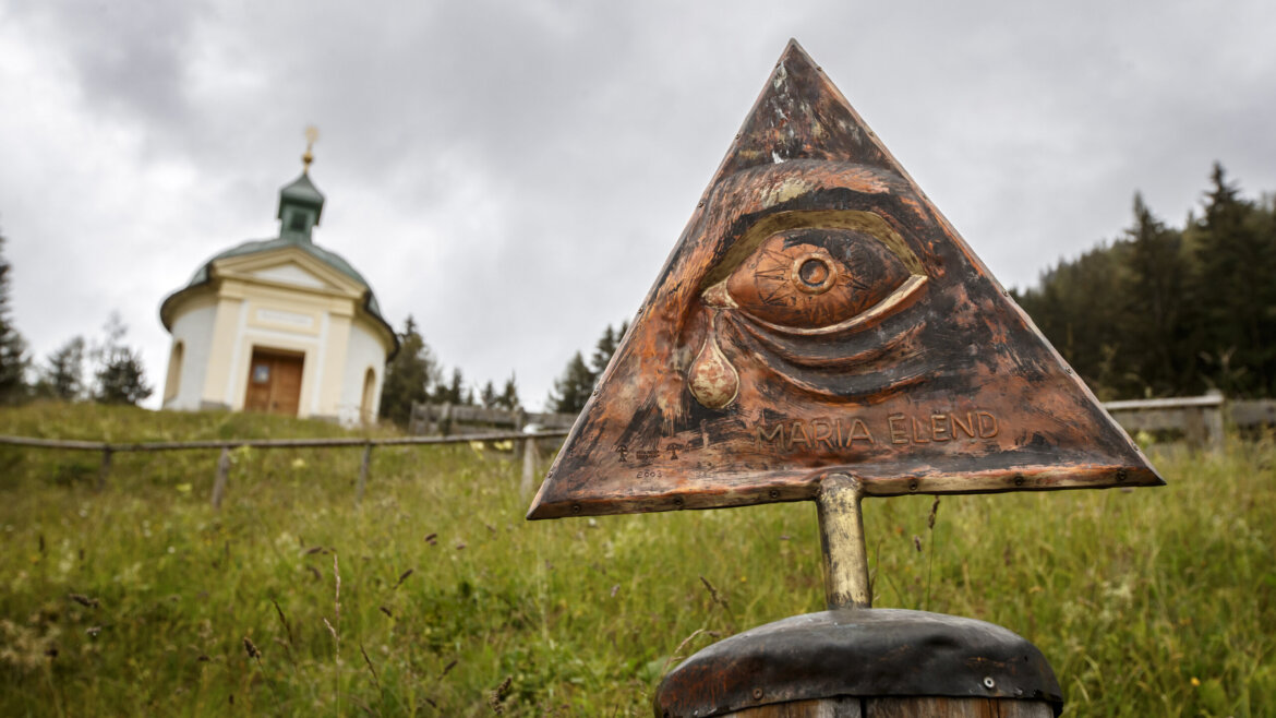 Eine dreieckige Metalltafel mit einem eingravierten Auge steht in der Nähe der Kapelle von Embach auf einem grasbewachsenen Hügel unter einem bewölkten Himmel. (vergrößerte Ansicht)