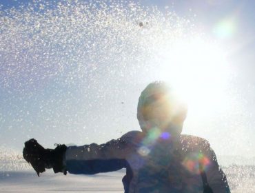 Kind in Winterkleidung wirft Schnee in die Luft auf einem sonnigen Eugendorfer Feld mit Bäumen im Hintergrund.