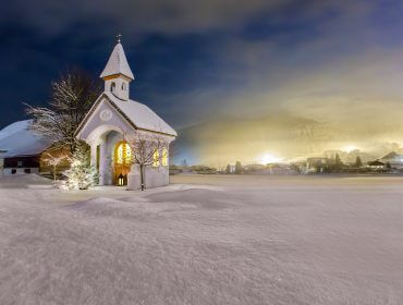 Eine kleine schneebedeckte Kirche leuchtet nachts in der Nähe von Hintersee in Österreich, umgeben von einer verschneiten Landschaft und fernen Dorflichtern.