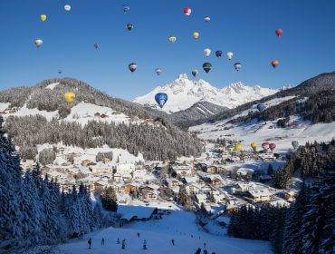 Bunte Heißluftballons schweben über Filzmoos, einem verschneiten österreichischen Bergdorf, mit Skifahrern auf einer Piste darunter.