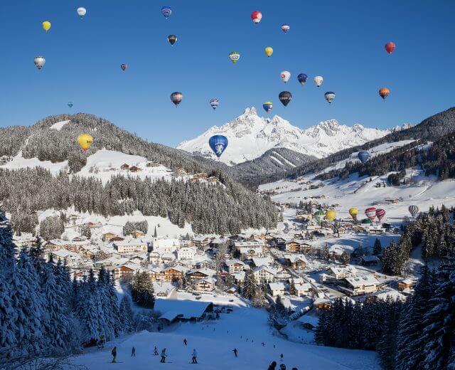Der Himmel über Filzmoos färbt sich bunt Bunte Heißluftballons schweben über Filzmoos, einem verschneiten österreichischen Bergdorf, mit Skifahrern auf einer Piste darunter.
