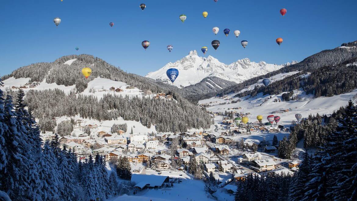 Heißluftballons schweben an einem sonnigen Tag über Filzmoos, einem österreichisch verschneiten Alpendorf mit Skifahrern und Bergen.