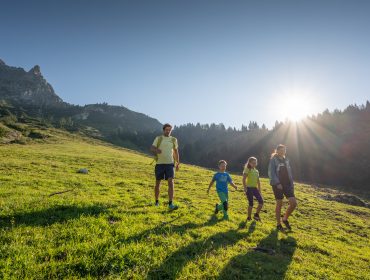 Eine vierköpfige Familie wandert über einen grasbewachsenen Filzmoos-Hang mit Bergen und Sonnenaufgang im Hintergrund.