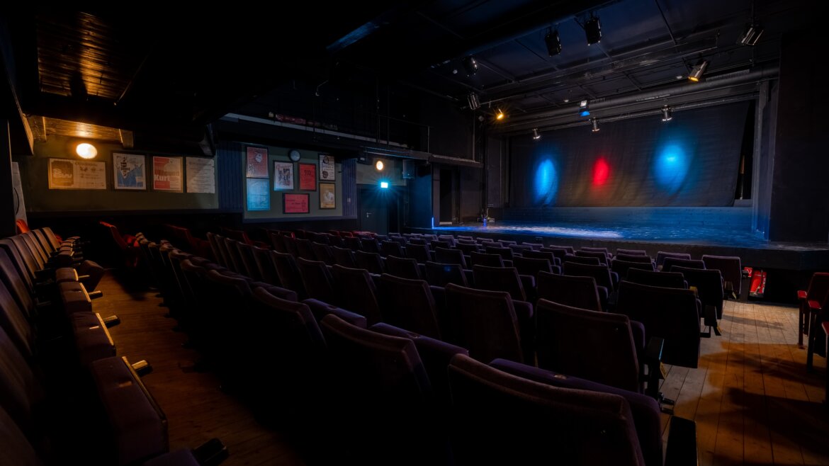 Leeres Cinetheatro in Neukirchen mit Sitzreihen vor einer von roten und blauen Scheinwerfern beleuchteten Bühne, Plakate an den Wänden. (vergrößerte Ansicht)