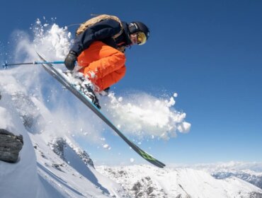 Skifahrer in orangefarbener Hose springt von einer verschneiten Piste im Gasteinertal, mit Bergen und blauem Himmel im Hintergrund.
