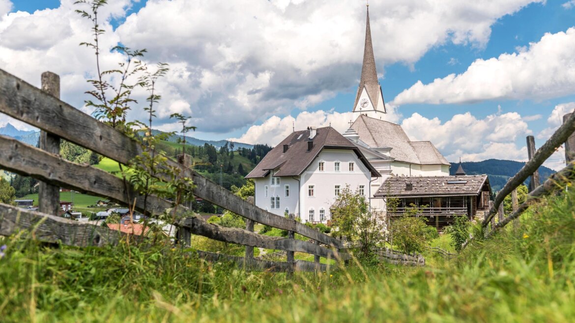 In Embach steht eine weiße Kirche mit einem hohen Kirchturm hinter einem Holzzaun in einer grasbewachsenen, ländlichen Landschaft. (vergrößerte Ansicht)