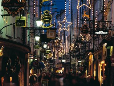 Getreidegasse im Advent – Christmasshopping im SL Eine festliche Straße in der Nacht, geschmückt mit Lichtersternen und Ladenschildern, in der sich die Menschen zum Weihnachtseinkauf tummeln.