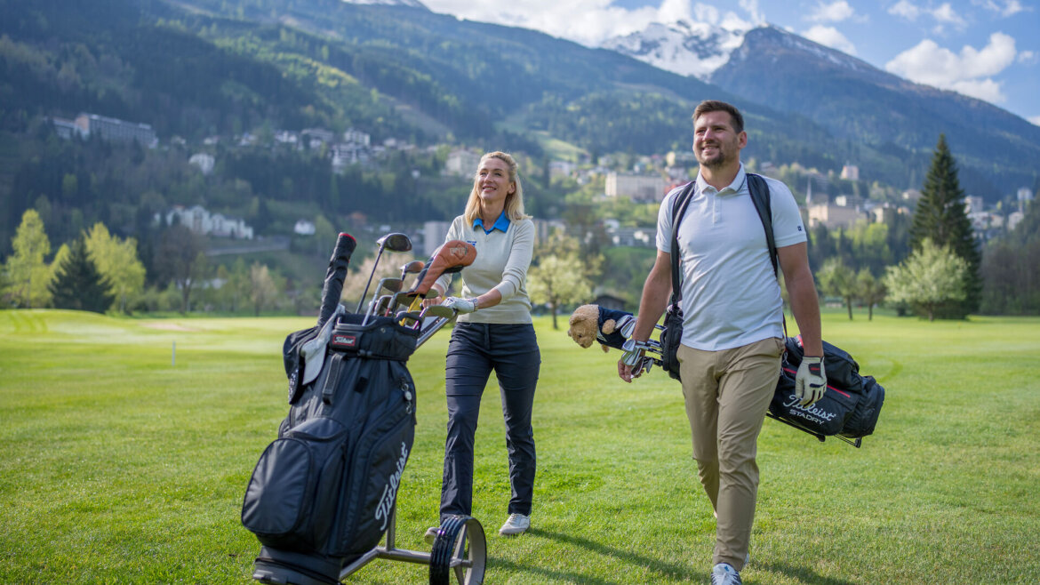 Zwei Personen gehen auf einem Gasteinertaler Golfplatz mit Schlägern, Lächeln, Bergen und Bäumen im Hintergrund. (vergrößerte Ansicht)