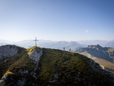 Zwei Wanderer wandern auf einem grasbewachsenen Bergrücken in der Nähe eines Gipfelkreuzes im Großarltal, mit fernen Gipfeln und blauem Himmel im Hintergrund.