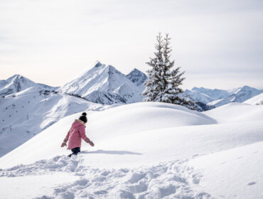 Schnee im Großarltal © Gipfelfieber Kind in rosa Mantel geht durch tiefen Schnee auf einem Hügel bei Großarl, mit verschneiten Bergen und Kiefern im Hintergrund.