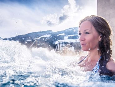 Alpine Wellness Frau in einem Freibad mit plätscherndem Wasser, schneebedeckten Bergen und Alpine Wellness im Hintergrund.