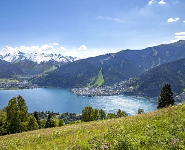 Der See ist umgeben von grünen Hügeln, der Stadt Zell am See und schneebedeckten Bergen unter einem blauen Himmel mit vereinzelten Wolken.