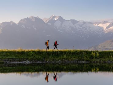 Zwei Wanderer mit Rucksäcken wandern entlang eines ruhigen Sees auf malerischen Weitwanderwegen, hinter denen sich schneebedeckte Gipfel erheben.