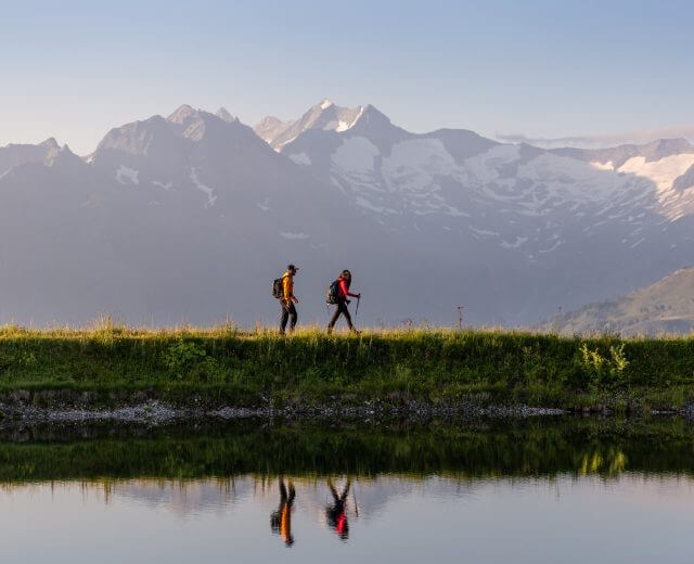 Zwei Wanderer mit Rucksäcken wandern entlang eines ruhigen Sees auf malerischen Weitwanderwegen, hinter denen sich schneebedeckte Gipfel erheben.
