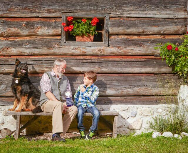 Ein Hund, ein älterer Mann und ein Bub sitzen auf einer Bank vor einer Holzhütte