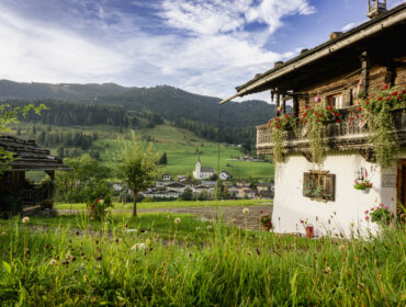 Alpendorf Embach mit grünen Hügeln, einer Kirche und einem blumengeschmückten Haus unter blauem Himmel.