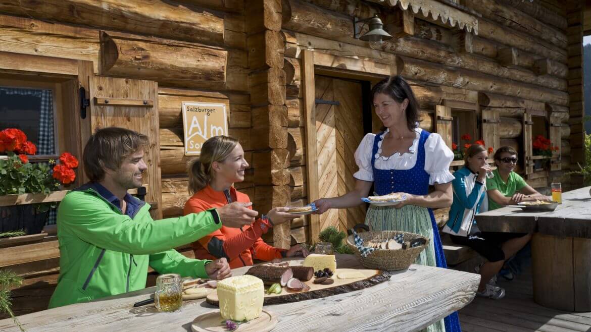 Paar sitzt vor einer Hütte und wird mit einer Jause bewirtet
