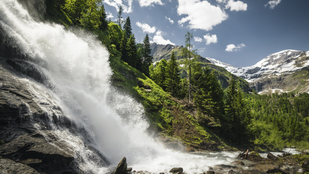 Großglockner Hochalpen Straße (c) Ferienregion Nationalpark hohe Tauern - Max Draege Mächtige Wasserfälle stürzen neben üppigen Wäldern in der Ferienregion Nationalpark Hohe Tauern in die Tiefe, dahinter schneebedeckte Gipfel. (vergrößerte Ansicht)