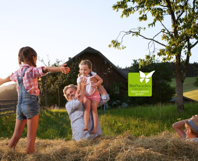 Eine Familie spielt auf einer sonnigen Wiese beim Bioparadies Biobauernhof, umgeben von Heu, Bäumen und Natur.