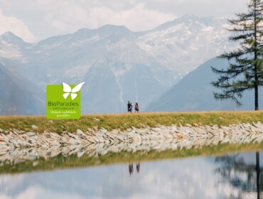 Zwei Personen wandern an einem Bergsee in der Nähe eines Biobauernhofs; Kiefern und Berge erheben sich unter einem bewölkten Himmel.