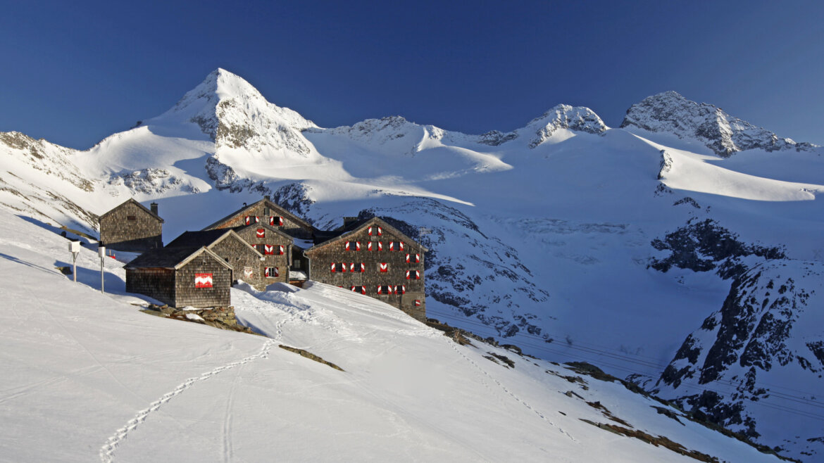 Steinerne Berghütten mit roten Fensterläden blicken in Neukirchen auf verschneite Hänge unter hohen Alpengipfeln. (vergrößerte Ansicht)