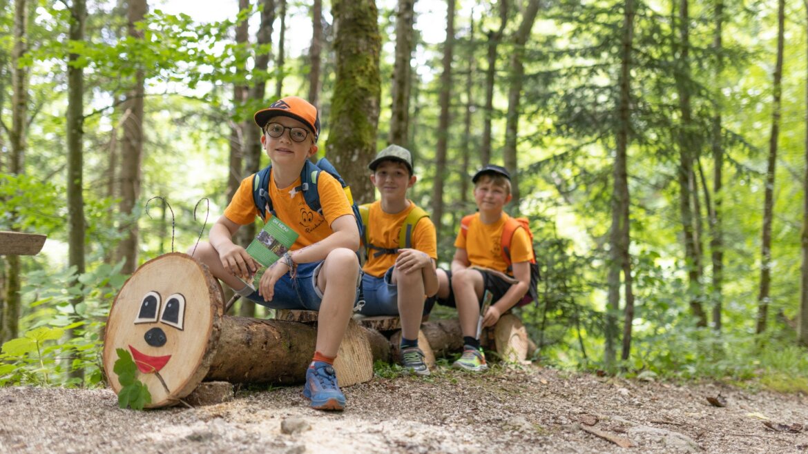 Drei Jungen in orangefarbenen Hemden sitzen auf einer Baumstamm mit einem Smiley-Gesicht im sonnigen Geisterwald in Russbach. (vergrößerte Ansicht)