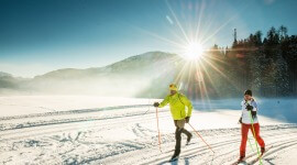 Zwei Personen beim Skilanglauf auf einer verschneiten Faistenauer Loipe bei strahlendem Sonnenschein und Bergen im Hintergrund.