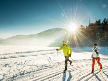 Zwei Personen beim Skilanglauf auf einer verschneiten Faistenauer Loipe bei strahlendem Sonnenschein und Bergen im Hintergrund.