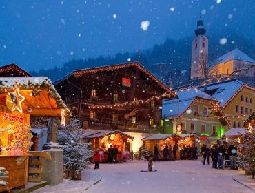 Festlicher Weihnachtsmarkt auf dem verschneiten Dorfplatz von Großarltal in der Abenddämmerung, mit Lichtern und geschmückten Holzbuden.