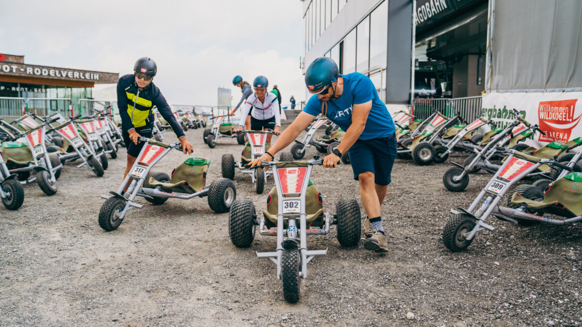 Helmträger bereiten sich an einem bewölkten Tag auf die Fahrt mit Bramberger Dreirädern auf einem Parkplatz vor, der mit ähnlichen Dreirädern gefüllt ist. (vergrößerte Ansicht)