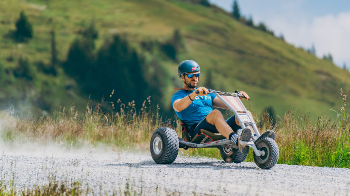 Ein Mann fährt mit einem dreirädrigen Go-Kart auf einem Schotterweg mit den grasbewachsenen Hügeln von Bramberg im Hintergrund. (vergrößerte Ansicht)