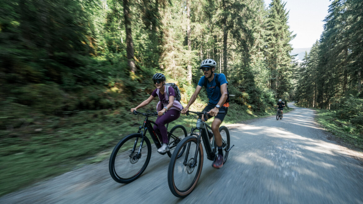 Zwei Personen fahren mit ihren Fahrrädern auf einem Waldweg in Bramberg, tragen Helme und sind von hohen Bäumen umgeben. (vergrößerte Ansicht)