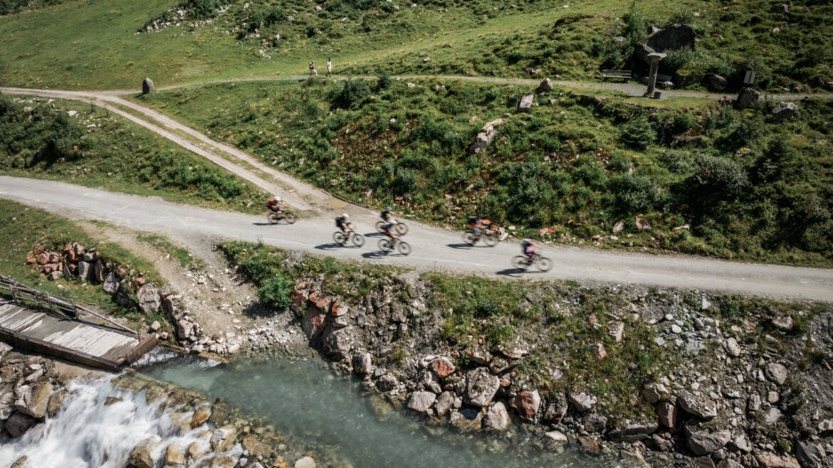Vier Radfahrer fahren auf einem unbefestigten Weg an einem felsigen Bach in der Nähe von Bramberg in einer grünen, bergigen Landschaft und neben einem Gebirgsbach (vergrößerte Ansicht)