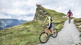 Mountainbik-(c)-Gasteinerta Zwei Radfahrer fahren auf einem Schotterweg durch die grüne Hügellandschaft im Gasteinertal, mit Bergen und einem Aussichtspunkt im Hintergrund.