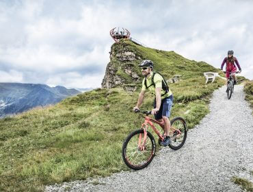 Zwei Radfahrer fahren auf einem Schotterweg durch die grüne Hügellandschaft im Gasteinertal, mit Bergen und einem Aussichtspunkt im Hintergrund.