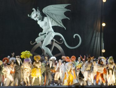 Eine große Gruppe in bunten Kostümen tritt bei den Salzburger Festspielen vor einer riesigen geflügelten Teufelsstatue auf.