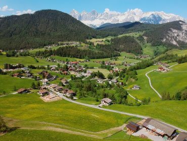 Luftaufnahme des grünen Tals von Forstau mit verstreuten Häusern, Hügeln und verschneiten Bergen unter blauem Himmel.