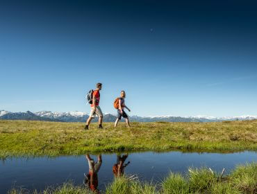 Zwei Personen wandern in der Nähe von Radstadt, Berge und blauer Himmel spiegeln sich im ruhigen Wasser.