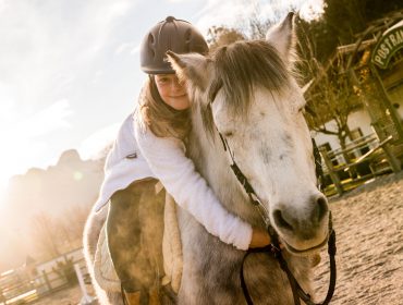 Ein Mädchen sitzt auf einem weiß-grauen Pferd und schlingt die Arme um dessen Hals.