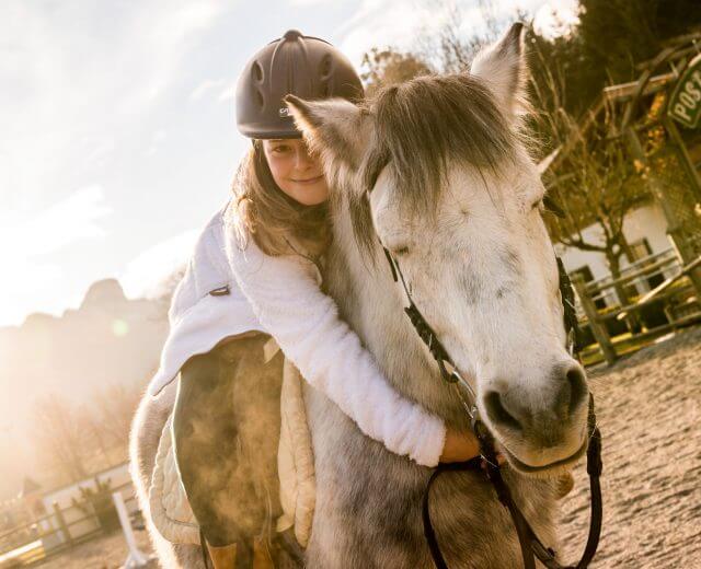Ein Mädchen sitzt auf einem weiß-grauen Pferd und schlingt die Arme um dessen Hals.
