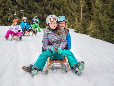 Zwei Mädchen in Winterkleidung genießen das Rodeln im Schnee, während andere Kinder in der Nähe zwischen verschneiten Bäumen Schlitten fahren.