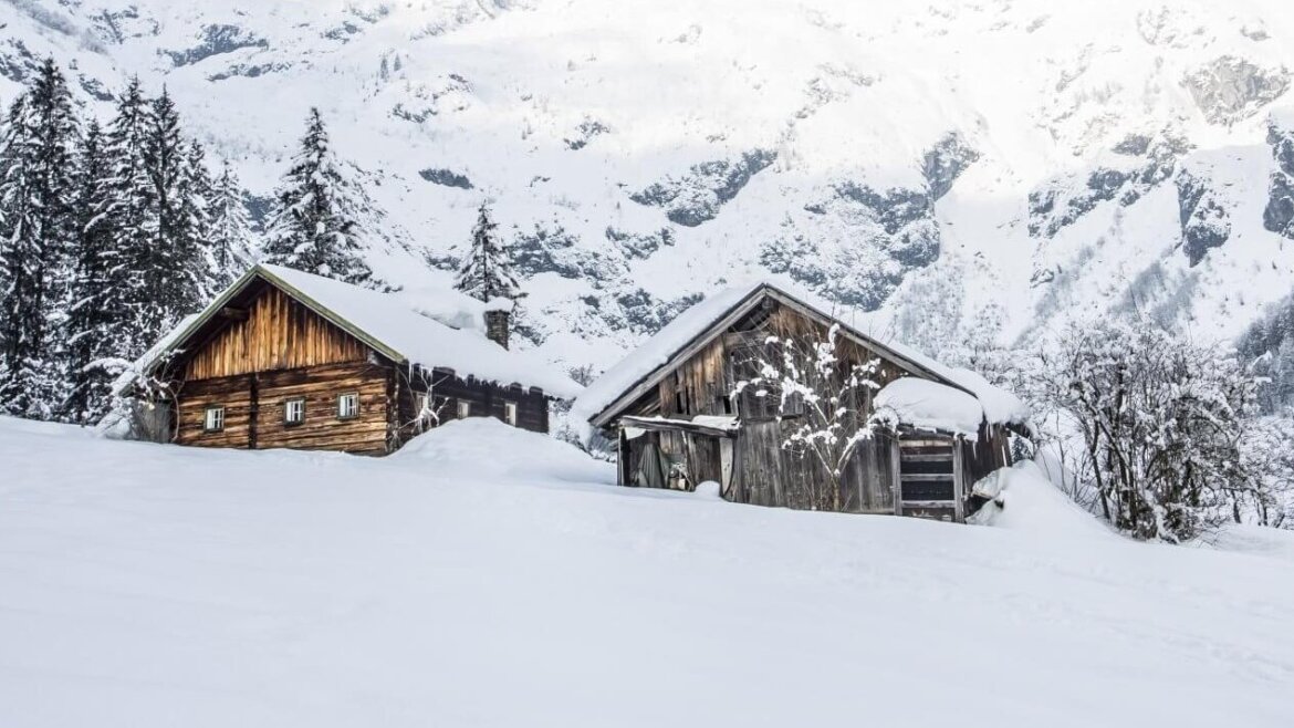 Zwei Holzhütten für einen Hüttenurlaub, eingebettet zwischen verschneiten Kiefern mit Bergen im Hintergrund. (vergrößerte Ansicht)