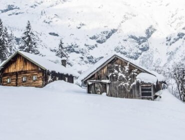 Zwei Holzhütten für einen Hüttenurlaub, eingebettet zwischen verschneiten Kiefern mit Bergen im Hintergrund.