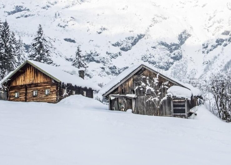 Winterlandschaft Zwei Holzhütten für einen Hüttenurlaub, eingebettet zwischen verschneiten Kiefern mit Bergen im Hintergrund.