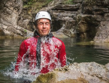 Mann mit Helm und Neoprenanzug taucht beim Canyoning in einer Schlucht aus dem Wildwasser auf