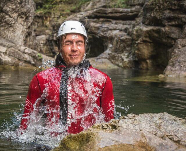 Mann mit Helm und Neoprenanzug taucht beim Canyoning in einer Schlucht aus dem Wildwasser auf