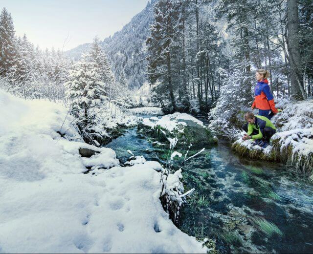 Zwei Läufer erfrischen sich am glasklaren Bach in einem verschneiten Wald