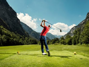 Ein Golfer schwingt seinen Schläger auf einem grünen Golfplatz, der von Bergen umgeben ist, unter einem blauen, teilweise bewölkten Himmel.